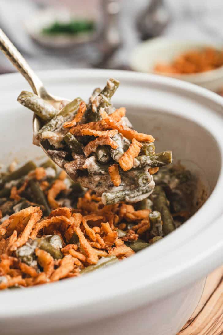 A spoon lifting a scoop of green bean casserole in a crockpot from the white bowl.