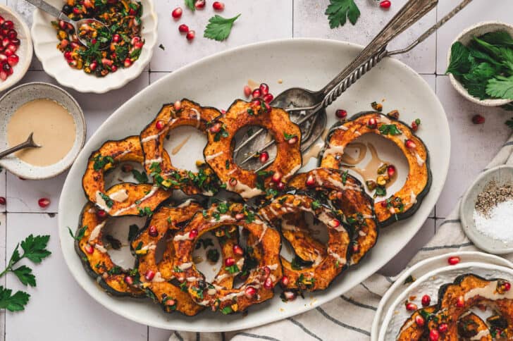 A white oval platter topped with oven roasted acorn squash with a creamy drizzle and pistachios and pomegranate seeds sprinkled over the top.