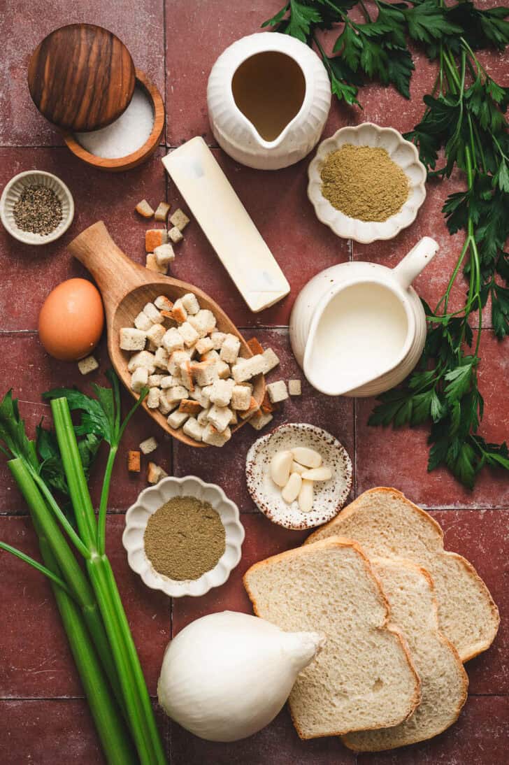 Ingredients on a dark red tiled surface, including bread and bread cubes, vegetables, an egg, butter, and herbs and spices.