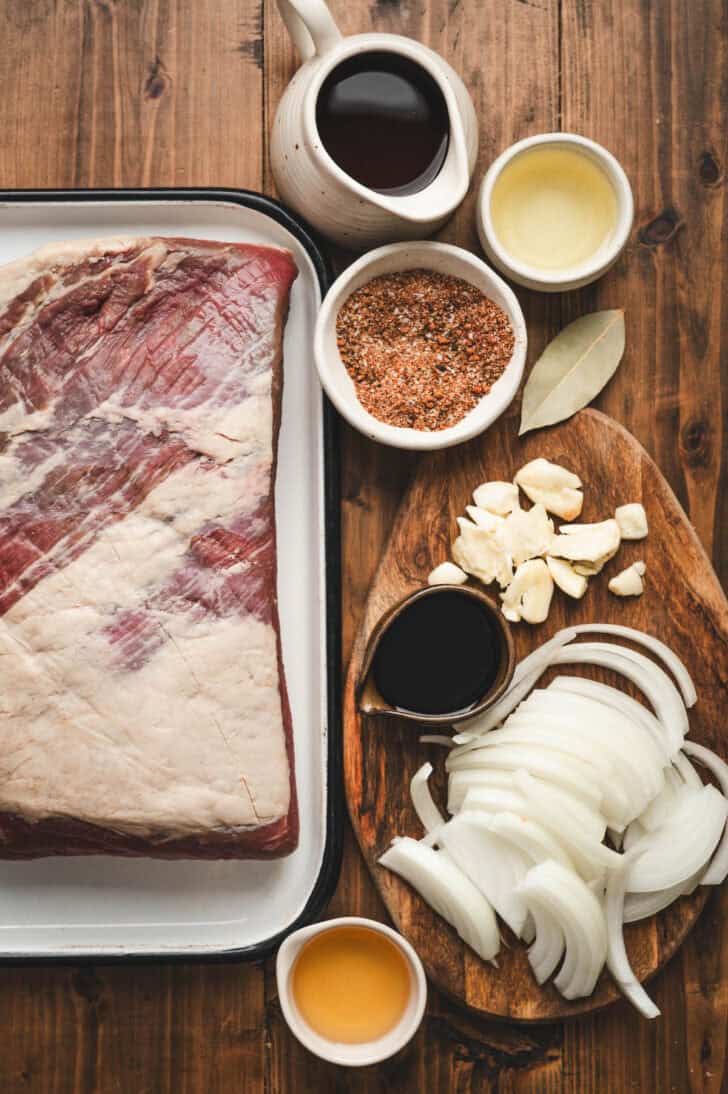 Ingredients on a wooden surface, including raw beef, spices, onion, garlic and condiments.