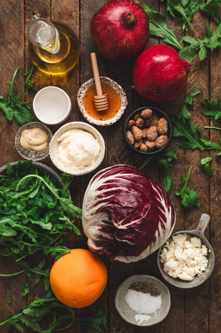 Ingredients on a wooden surface, including arugula, radicchio, oranges, nuts, pomegranate, mayo, vinegar and cheese.