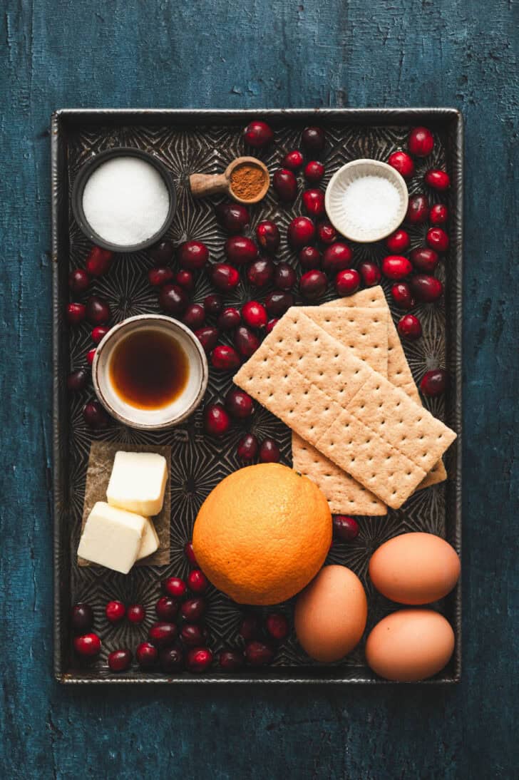Ingredients on a baking pan, including graham crackers, orange, eggs, sugar, butter and berries.