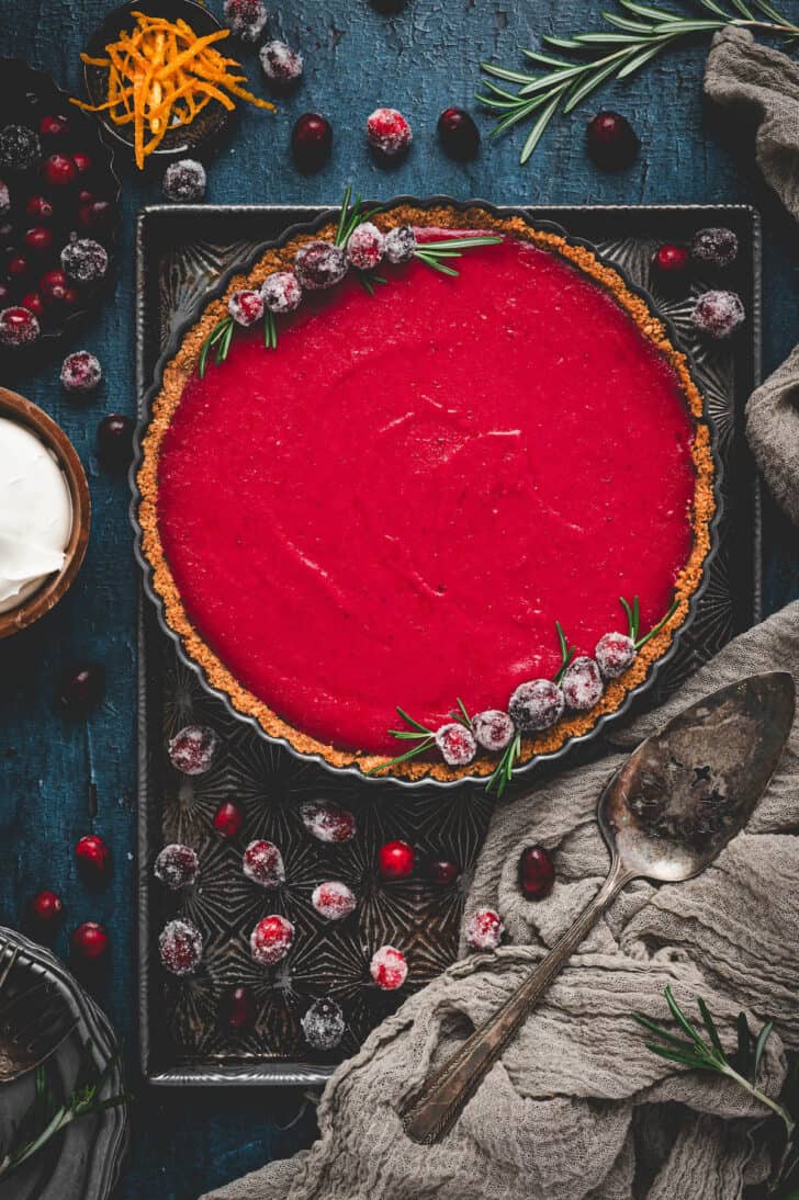 A cranberry curd tart garnished with sugared cranberries on a textured baking pan.