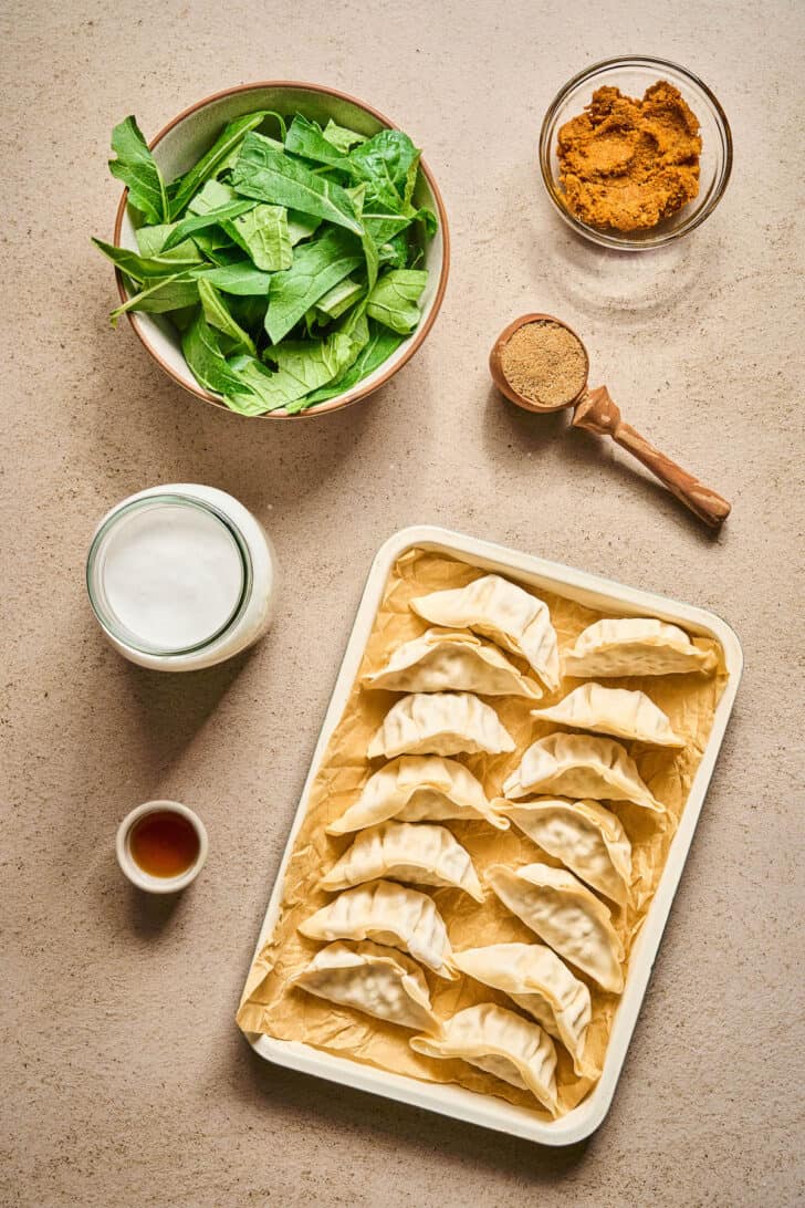 Ingredients on a tan background, including potstickers, chopped greens, coconut milk, fish sauce and spices.