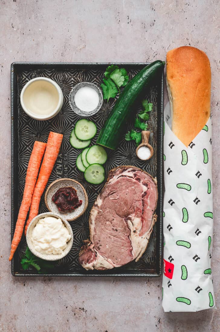 Ingredients on a light surface including a large piece of cooked beef, cucumber, carrots, mayonnaise, hot sauce, herbs and Jimmy John's day old bread.