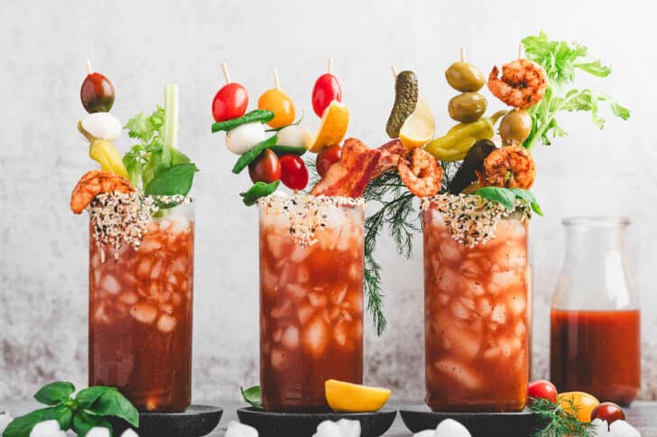 Three glasses filled with a red cocktail mix and garnishes on bloody mary skewers.