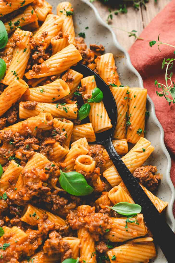 A platter filled with rigatoni bolognese with a black serving spoon in it.