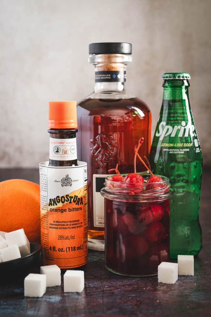 Ingredients in front of a light background, including bitters, a bottle of sprite, sugar cubes, cherries and a bottle of brown liquor.