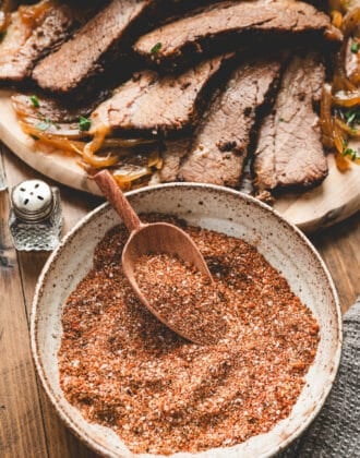 A bowl of brisket rub recipe with a small wooden scoop in it, with a plate of cooked sliced brisket in the background.