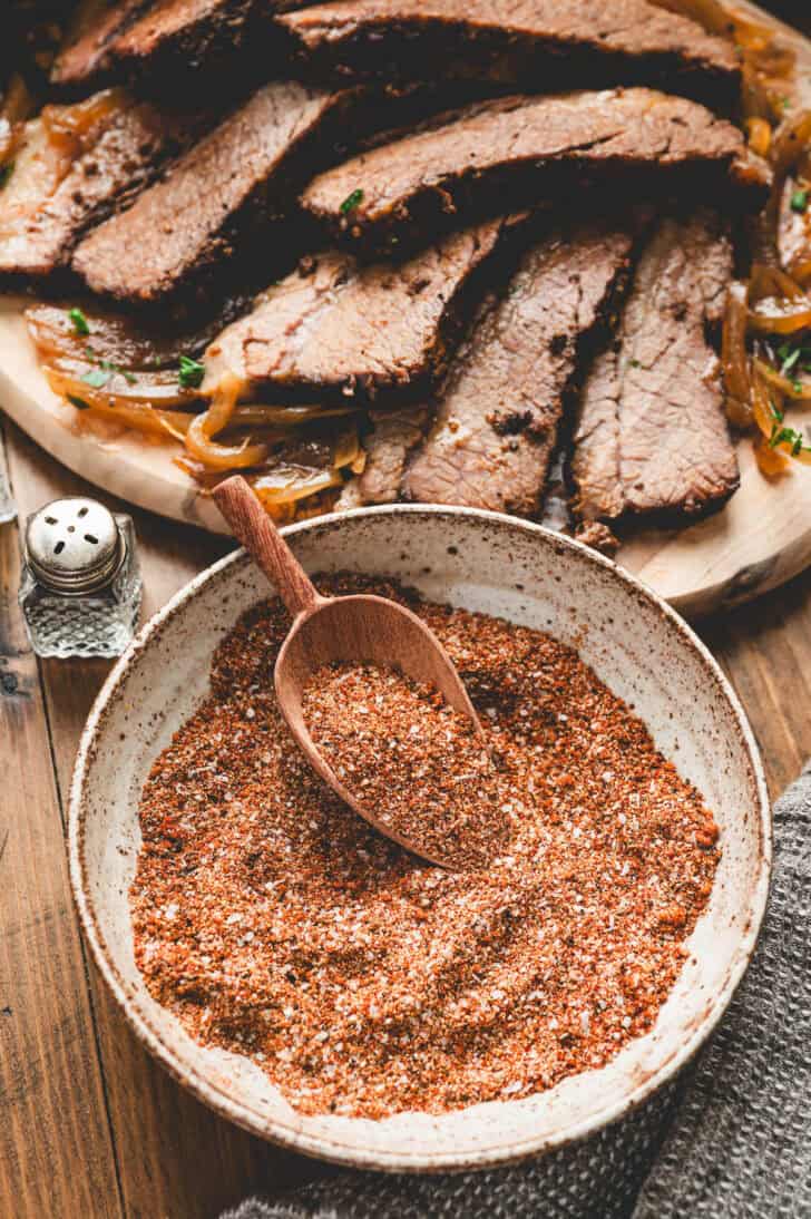 A bowl of brisket rub recipe with a small wooden scoop in it, with a plate of cooked sliced brisket in the background.