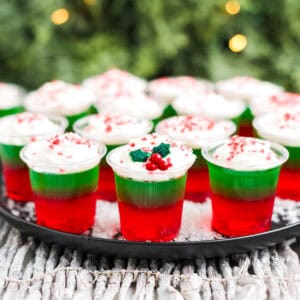 Christmas jello shots on a wooden cutting board topped with whipped cream and festive sprinkles.
