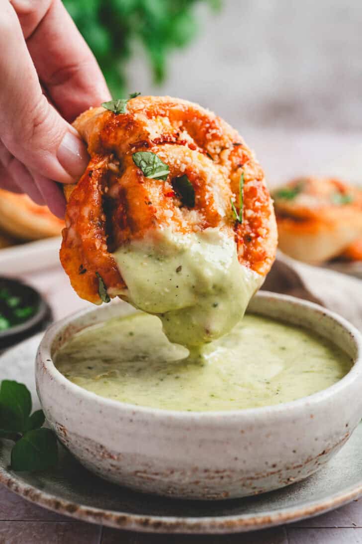 A hand dipping a pizza roll into a bowl of pesto ranch salad dressing.