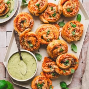 A pizza pinwheels recipe displayed on a geometric white platter with a green dipping sauce.
