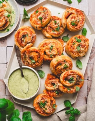 A pizza pinwheels recipe displayed on a geometric white platter with a green dipping sauce.