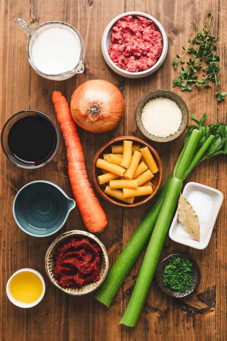 Ingredients on a wooden table, including pasta, carrots, celery, onion, ground beef, tomato paste, herbs and spices.