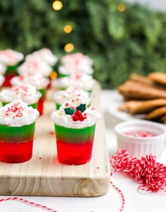 Christmas jello shots on a wooden cutting board topped with whipped cream and festive sprinkles.