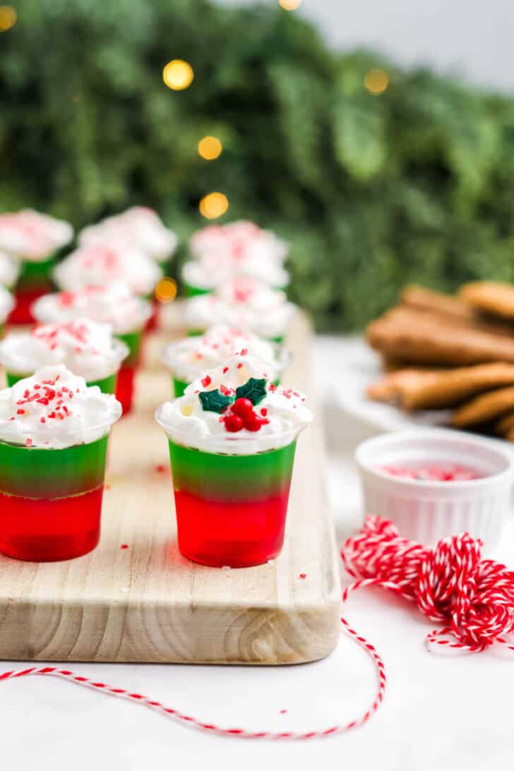 Christmas jello shots on a wooden cutting board topped with whipped cream and festive sprinkles.