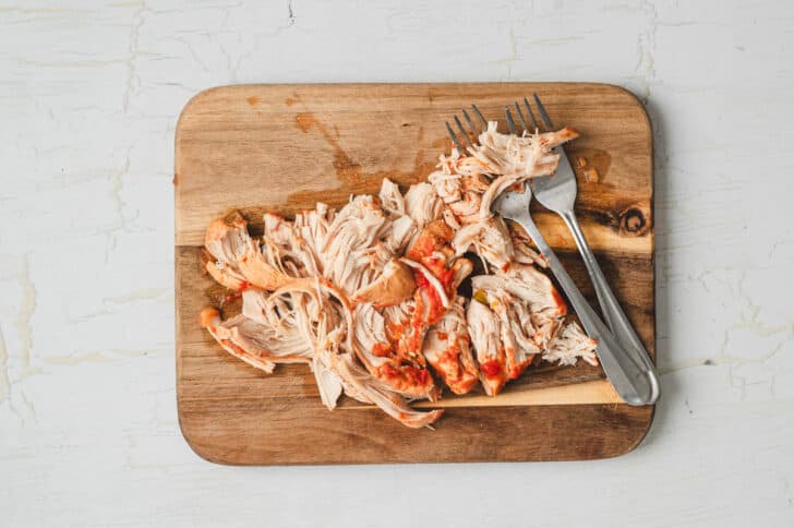 A cooked chicken breast being shredded with forks on a wooden cutting board.