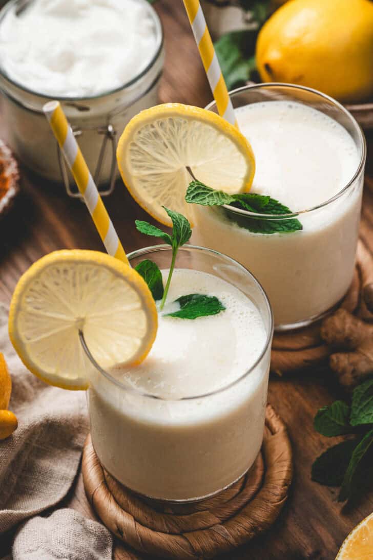 Two servings of lemon ginger smoothie garnished with a fresh lemon wheel, mint, and a yellow straw.