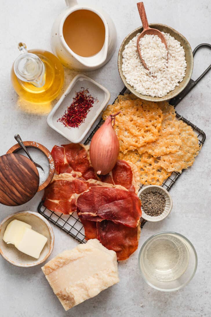 Ingredients on a light background including rice, saffron, butter, oil, spices, shallot, prosciutto and crispy parmesan.