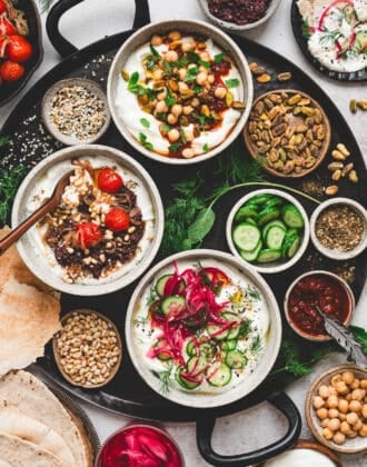 A black platter filled with three bowls of savory Greek yogurt recipes, along with various toppings for them.