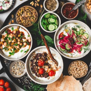A black platter filled with three bowls of savory Greek yogurt recipes, along with various toppings for them.