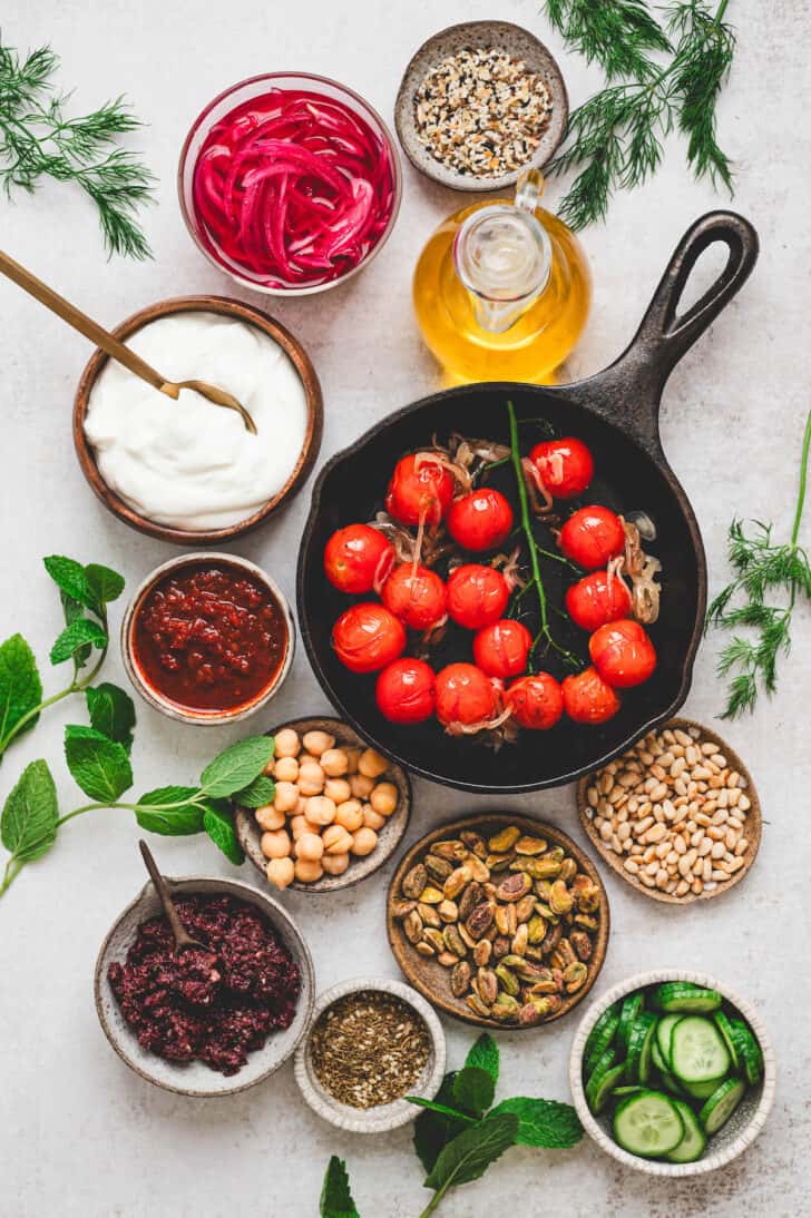 Ingredients on a light background, including blistered tomatoes, harissa, chickpeas, nuts, olive oil, pickled onions, herbs, spices, cucumbers and yogurt.