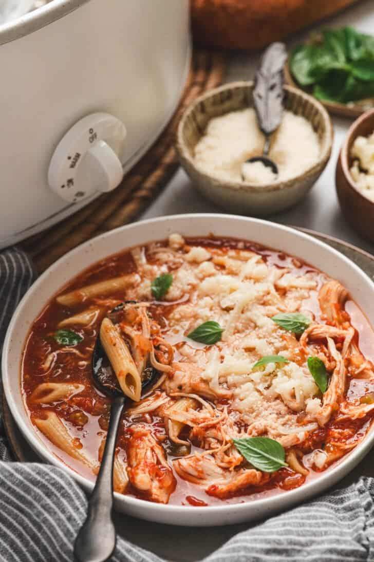 A bowl of a chicken parmesan soup recipe sitting in front of a crockpot.