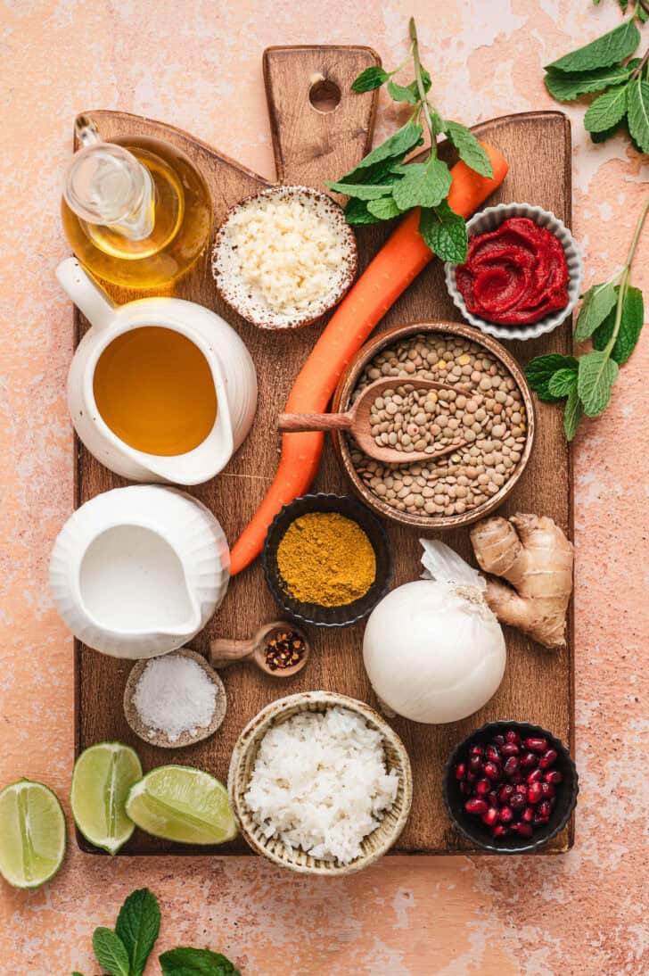 A wooden cutting board topped with ingredients, including dried legumes, carrot, garlic, lime, rice, broth, tomato paste, herbs and spices.