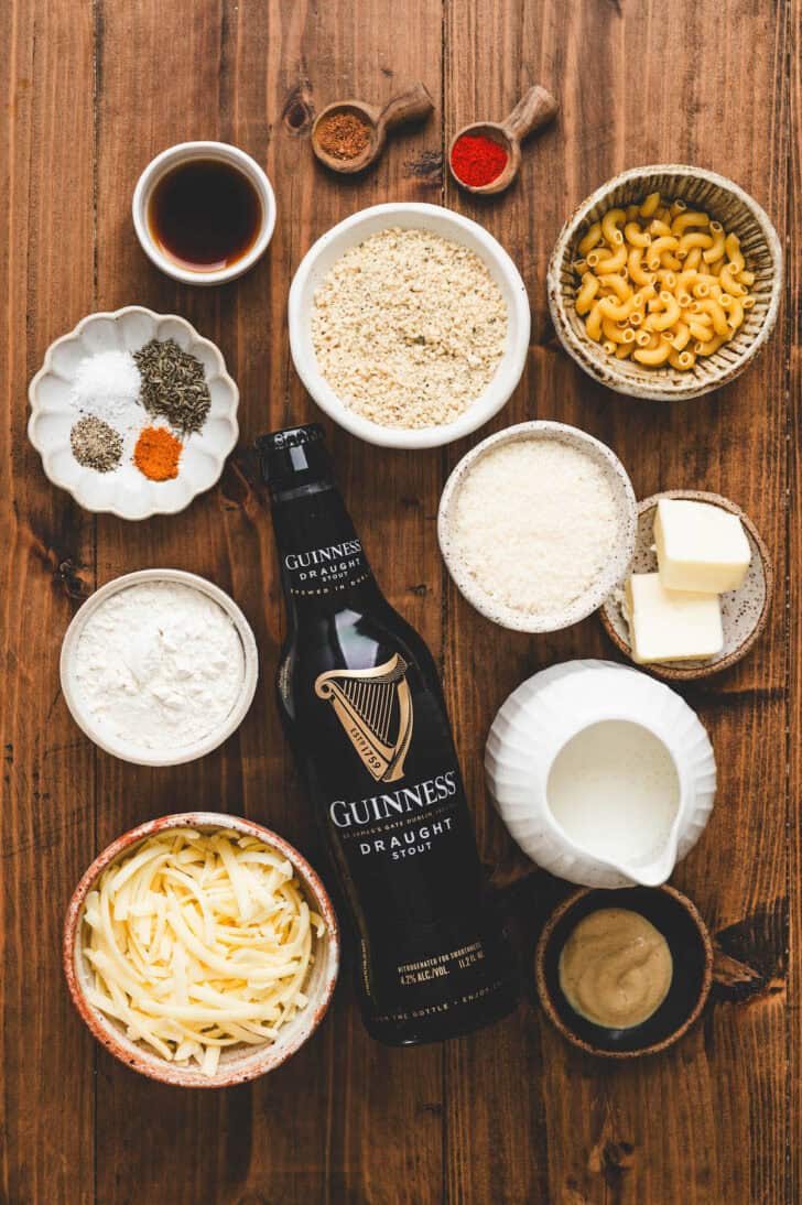 Ingredients on a wooden surface, including shredded cheese, elbow noodles, breadcrumbs, herbs and spices, milk and a bottle of Guinness.