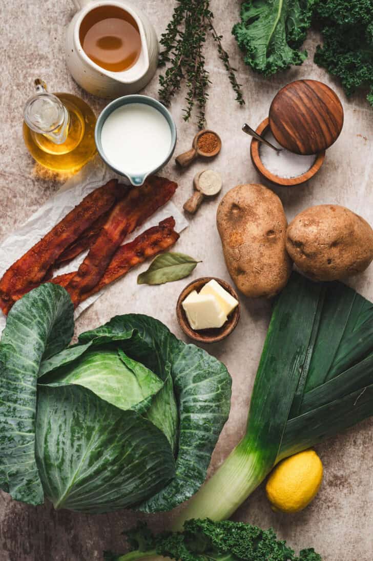 Ingredients on a light surface, including cabbage, kale, potatoes, butter, milk, oil, lemon, herbs and spices.