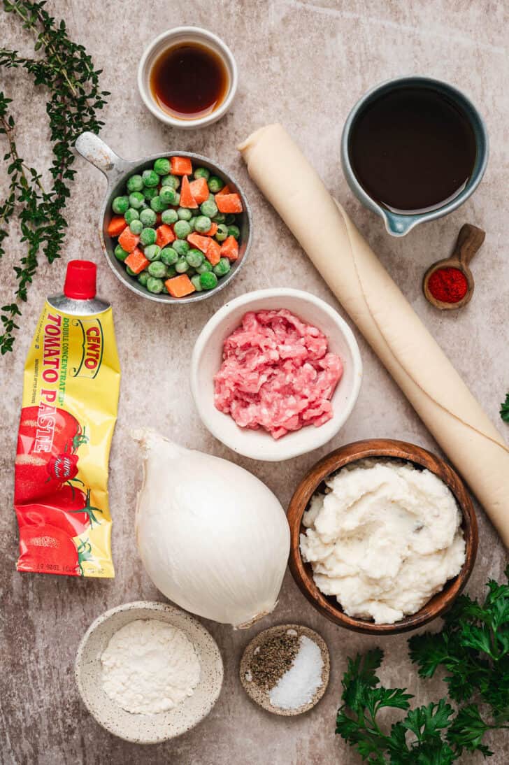 Ingredients on a light background, including ground meat, mashed potatoes, frozen peas and carrots, pie crust, onion, herbs and tomato paste.