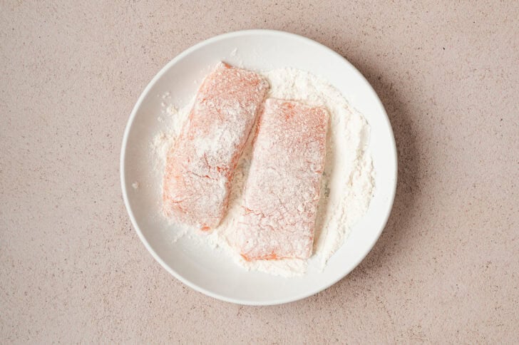 Two pieces of fish being dredged in flour in a white bowl.