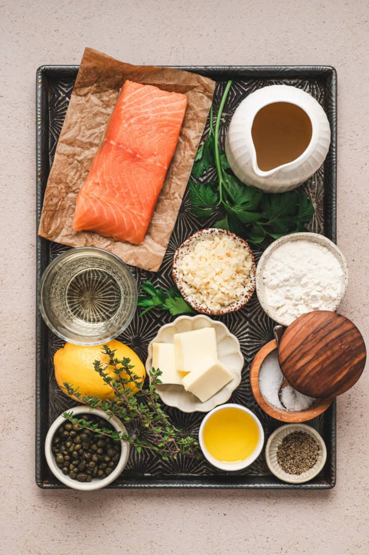 Ingredients on a baking pan, including raw pink fish, butter, lemon, herbs, flour, capers and oil.