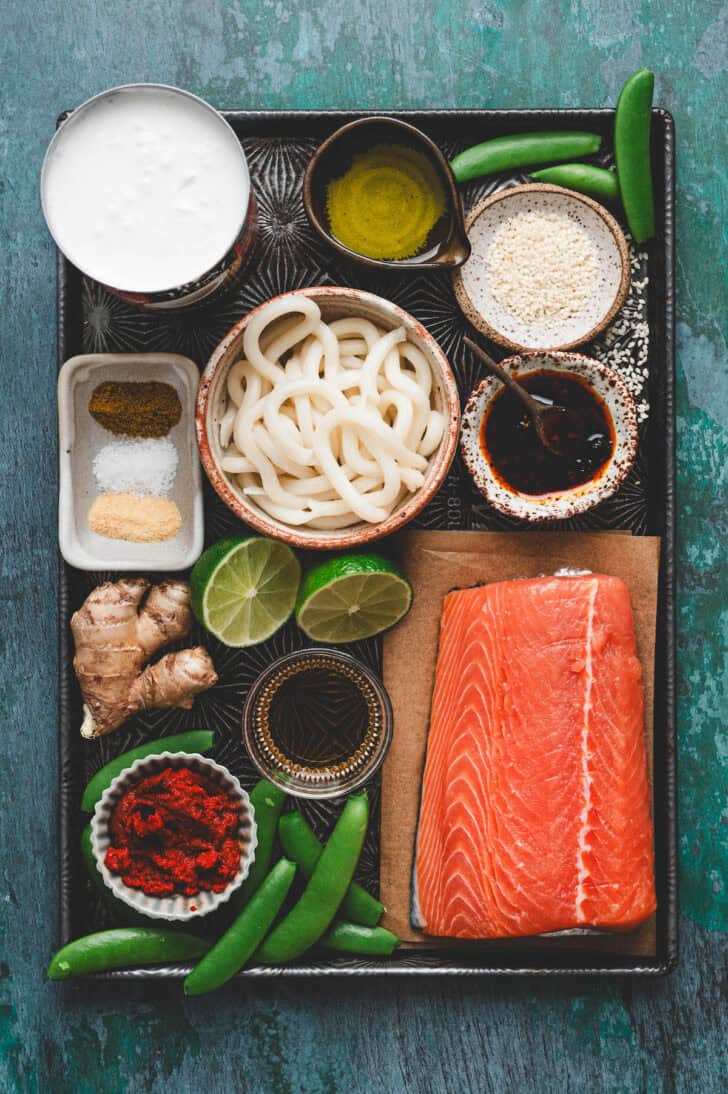 Ingredients on a baking pan, including raw fish, udon noodles, snap peas, lime, ginger and spices.