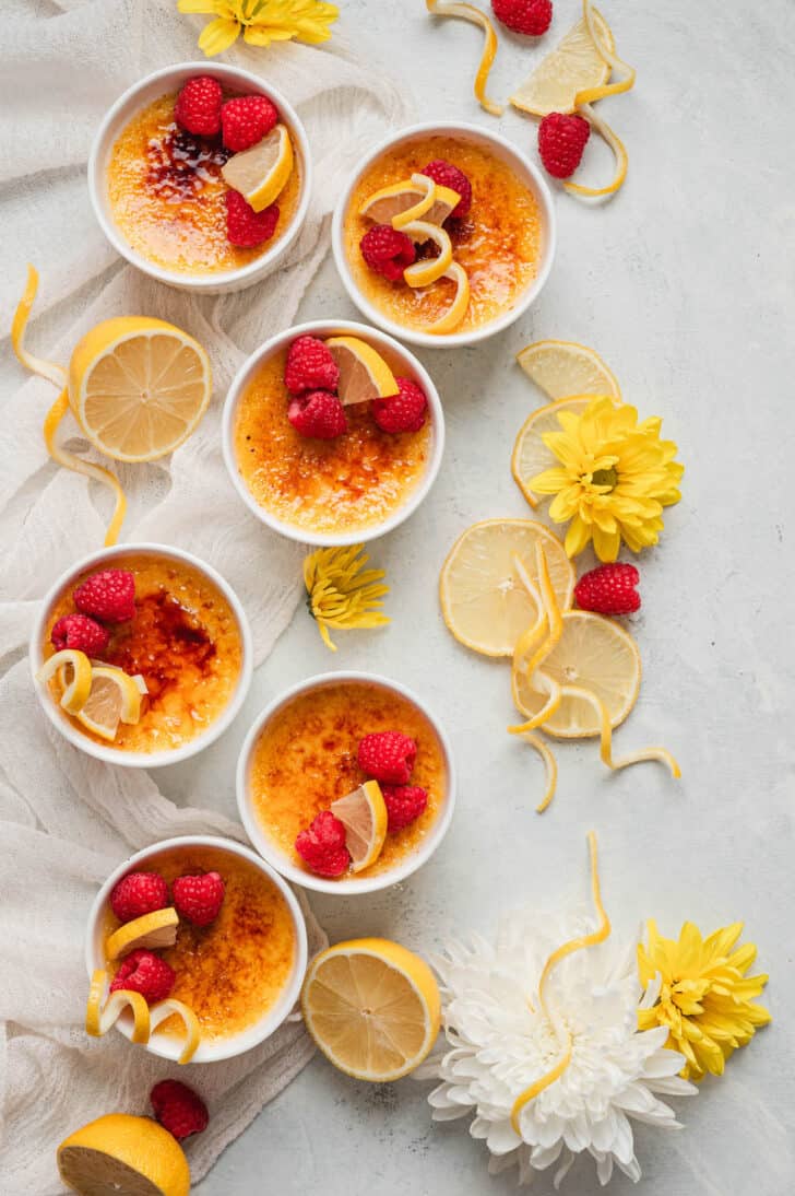 Six ramekins of lemon creme brulee garnished with raspberries and lemon peel, on a white surface with flowers decorating the scene.