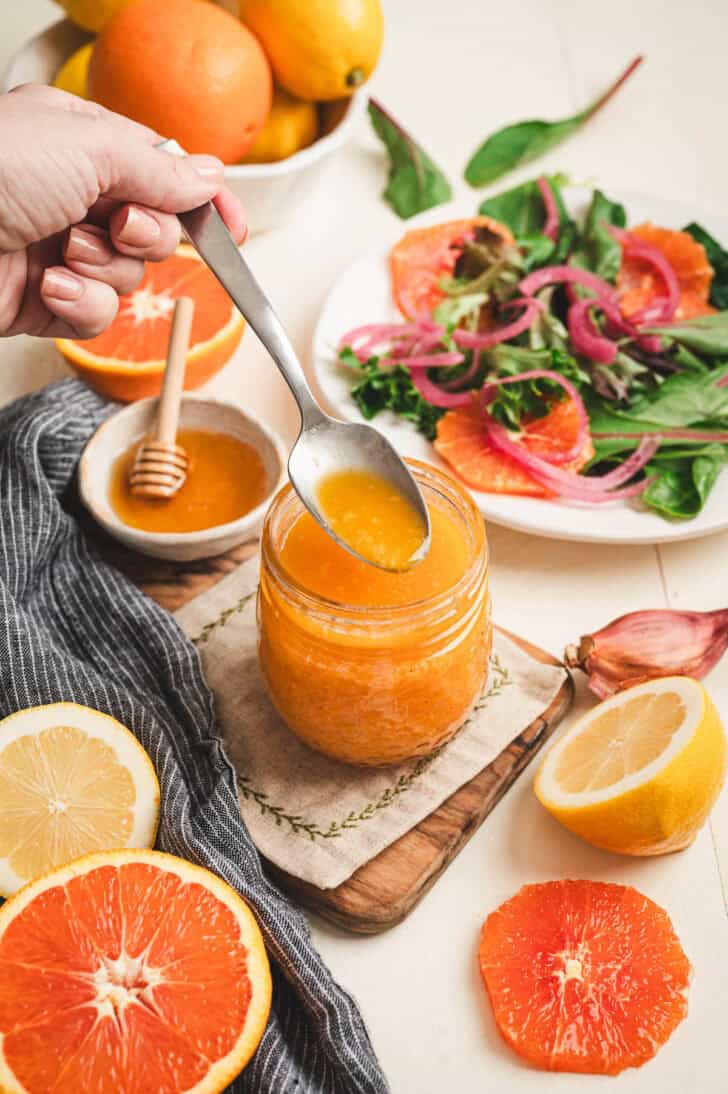 A spoon lifting some citrus vinaigrette from a glass jar with salad fixings in the background and foreground.