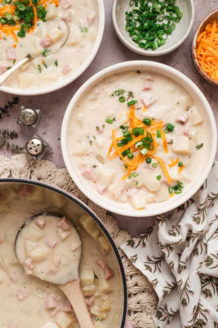 A bowl of potato ham soup alongside the pot with a wooden spoon in it.