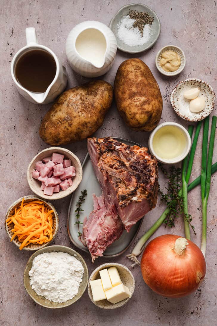 Ingredients on a neutral surface including potatoes, ham and a ham bone, onions, butter, cheese, flour, garlic and dairy products.