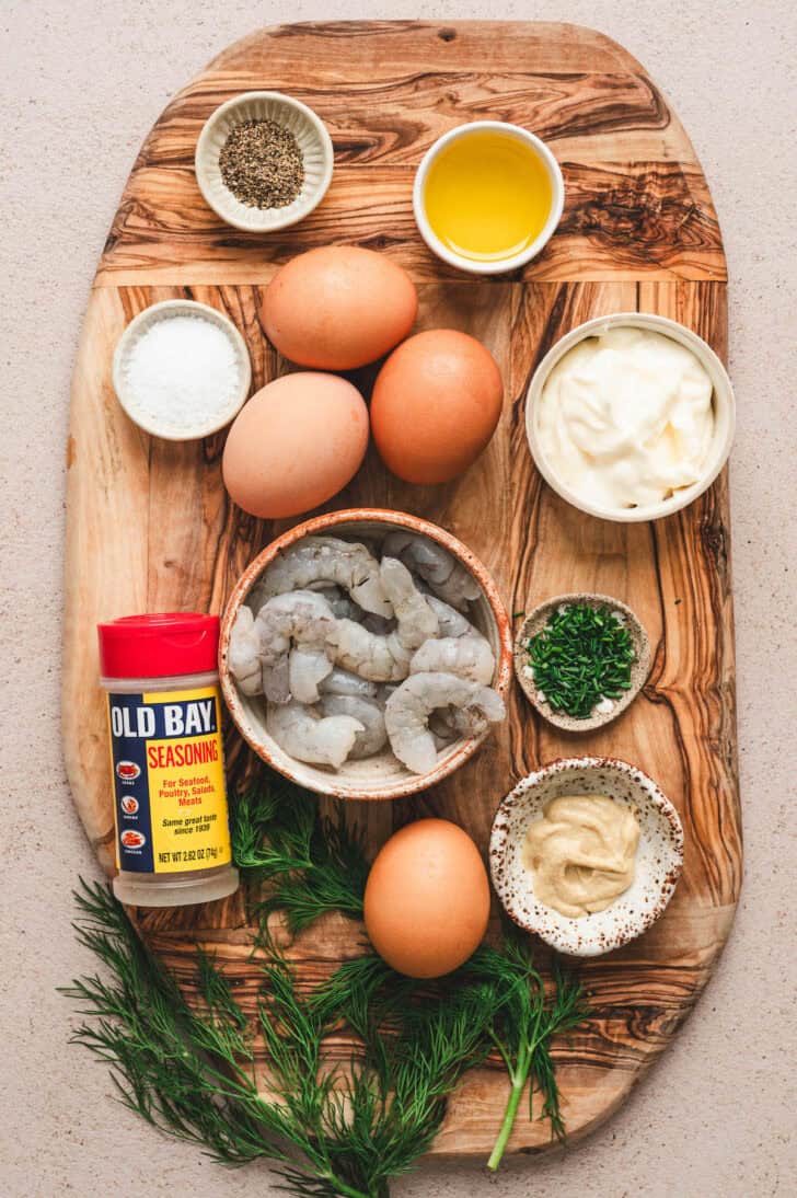 Ingredients on a wooden cutting board, including prawns, old bay, eggs, herbs and spices.
