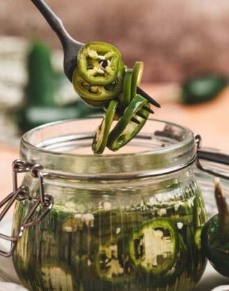A fork lifting pickled jalapeno peppers from a glass jar.