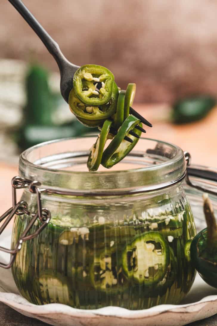 A fork lifting pickled jalapeno peppers from a glass jar.