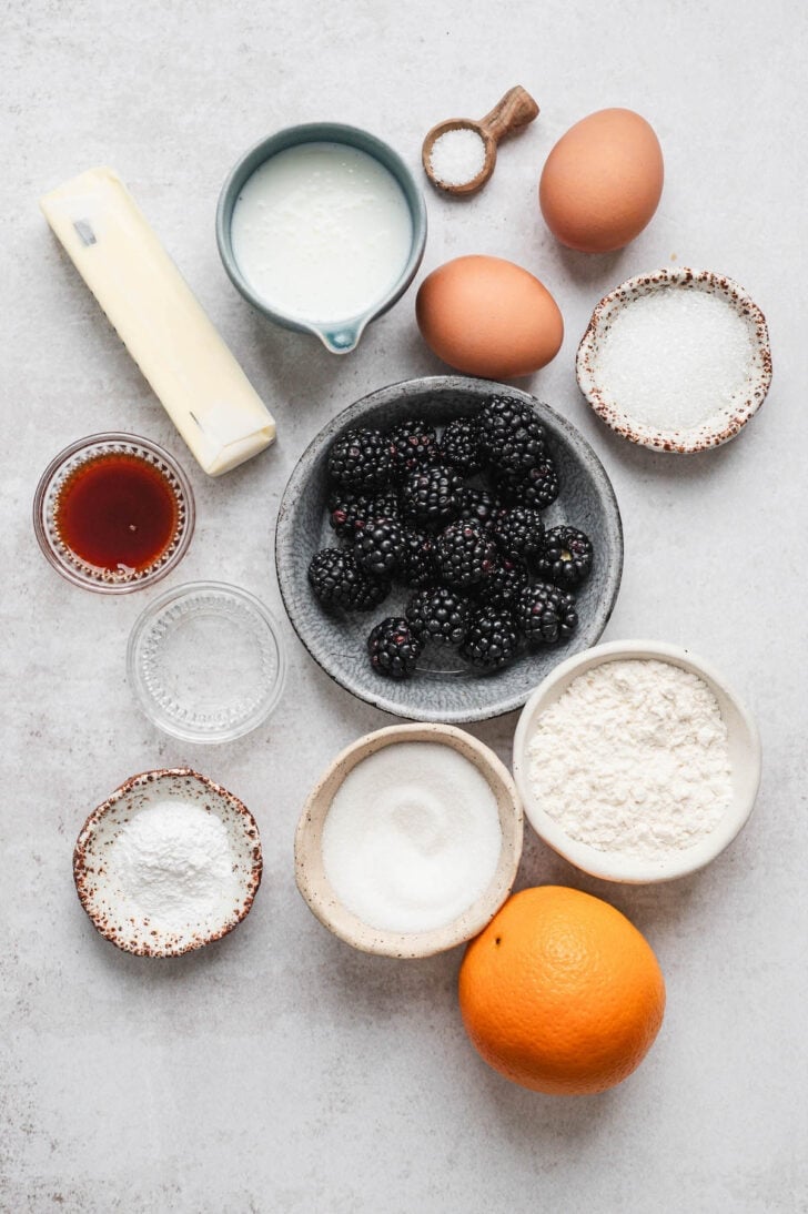 Ingredients on a light surface, including bowls with berries, flour, sugar, eggs, an orange, and butter.