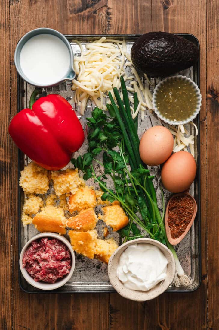 Ingredients on a baking pan on a wooden surface, including cornbread, eggs, green onions, bell peppers, cheese and dairy ingredients.