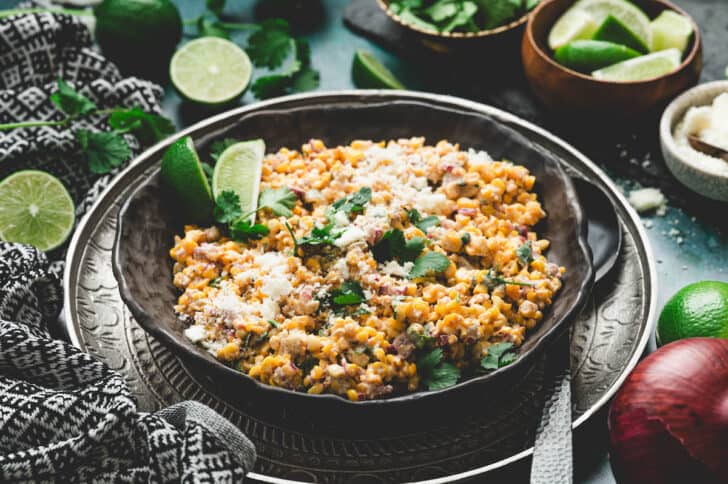 A bowl of elote corn salad garnished with crumbled cheese and lime wedges.