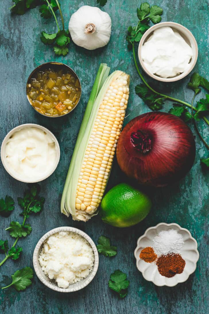 Ingredients on a teal surface, including corn on the cob, red onion, lime, green chiles, garlic, spices, cilantro and dairy products.