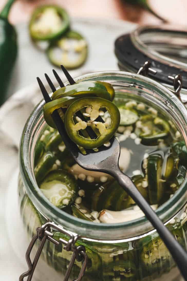 A fork lifting the best pickled jalapenos recipe from a glass jar.