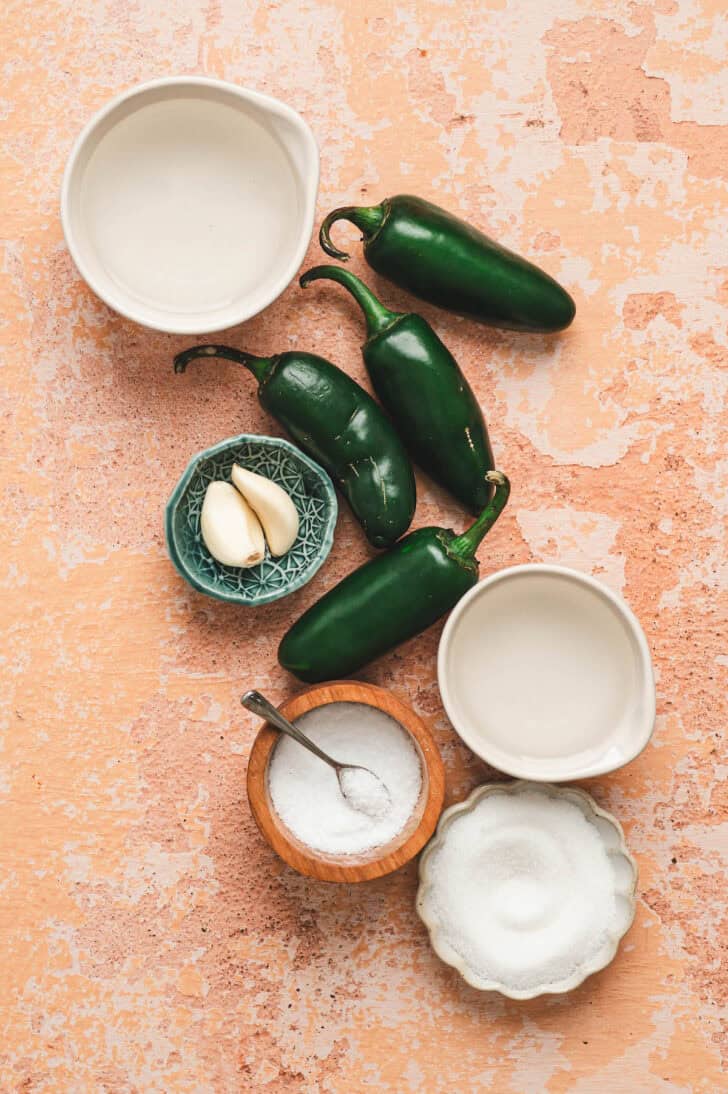 Ingredients on a light orange background, including jalapeno peppers, sugar, vinegar and garlic.