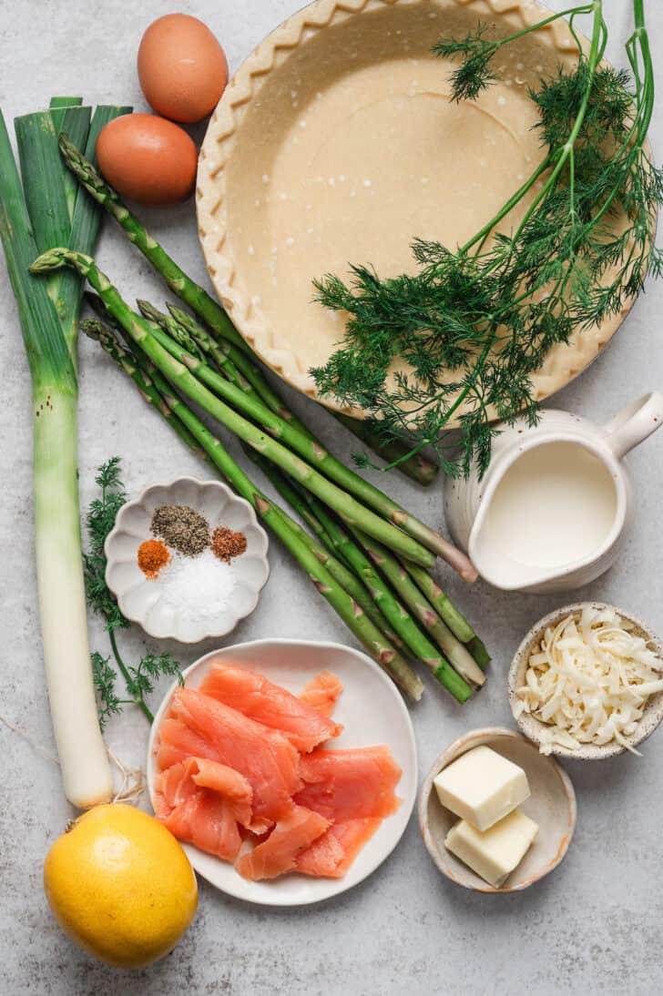 Ingredients on a light background, including a pie crust, smoked salmon, asparagus, eggs, leek, lemon, herbs and spices.