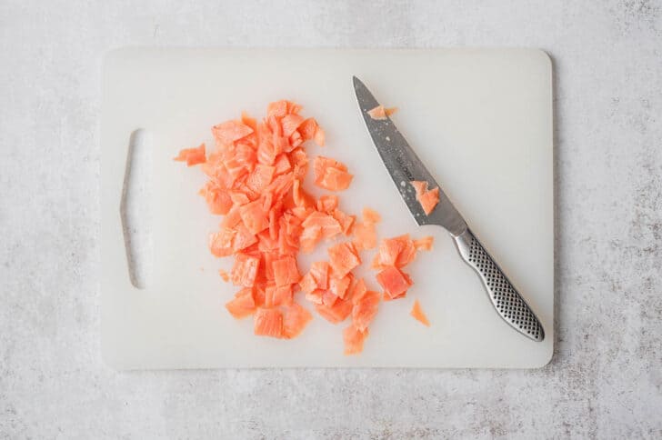 A knife chopping smoked salmon on a white cutting board.